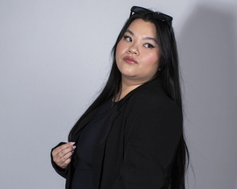 A young woman standing in front of a grey wall. Her body is facing the left side while her head is facing the camera. Her eyes are looking to the top right.